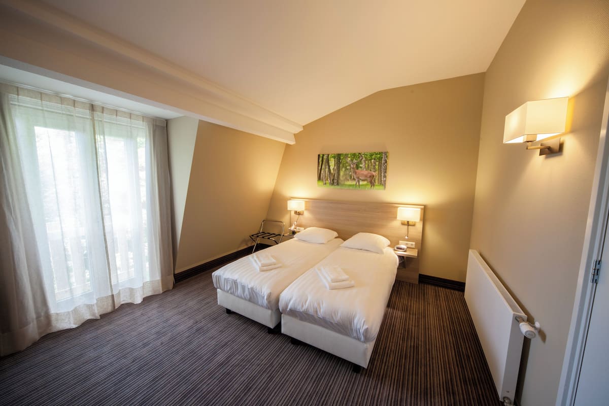 A hotel room with two beds, a large window with sheer curtains, a painting of a deer, and a radiator.