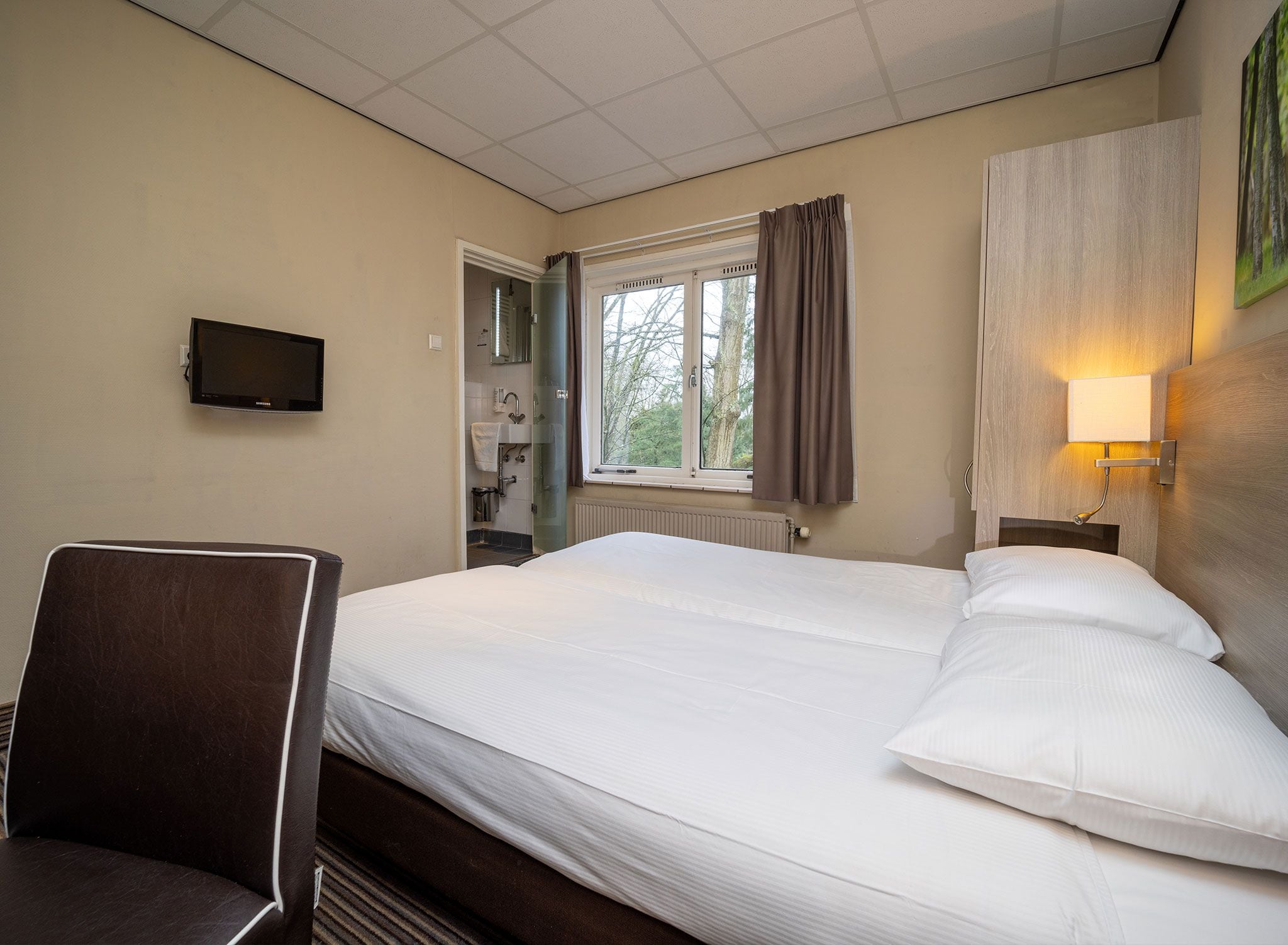 A hotel room with a bed, a desk, a television, a window with curtains, and a wall-mounted light fixture.