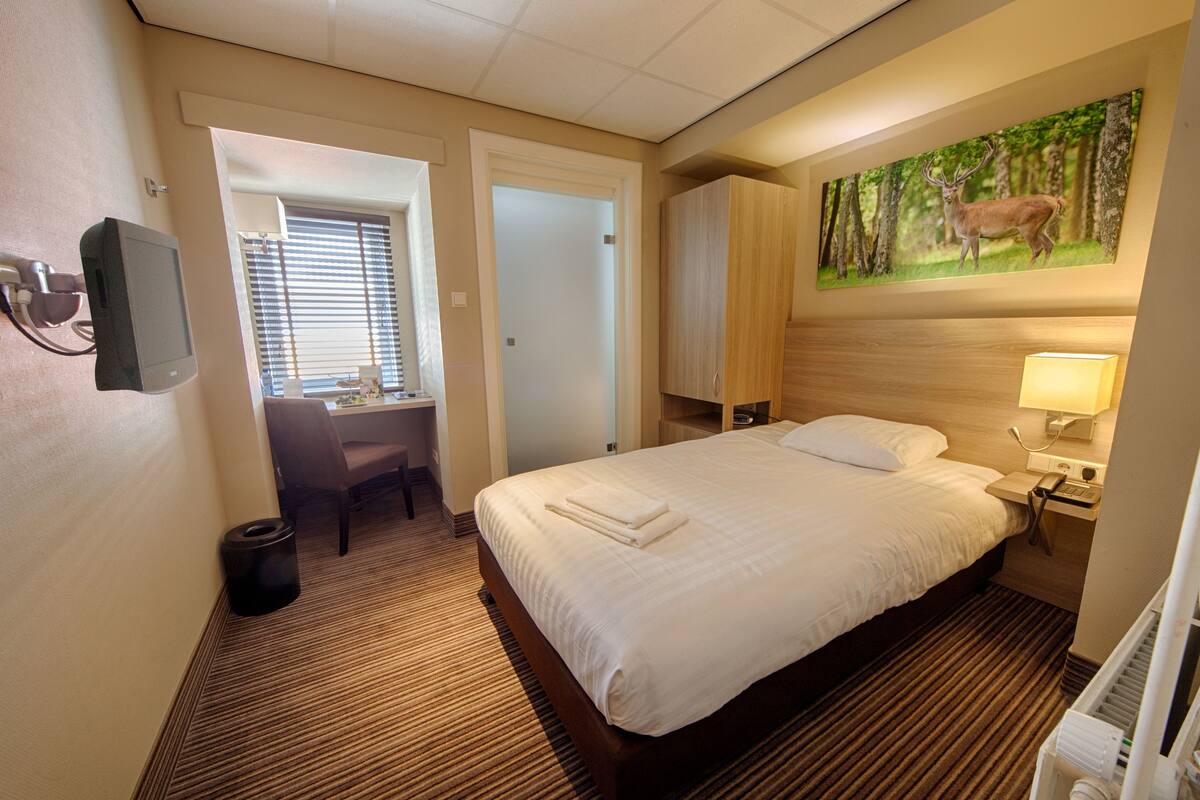 Een hotelkamer met een groot bed, een bureau met stoel, een schilderij van een hert, en een aan de muur bevestigde televisie.