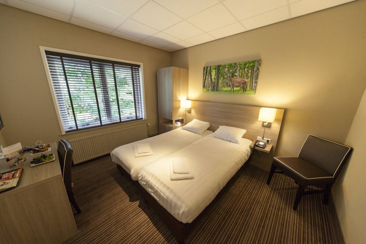 Een hotelkamer met een groot bed, twee nachtkastjes, een bureau met stoel, een radiator en een raam met jaloezieën.
