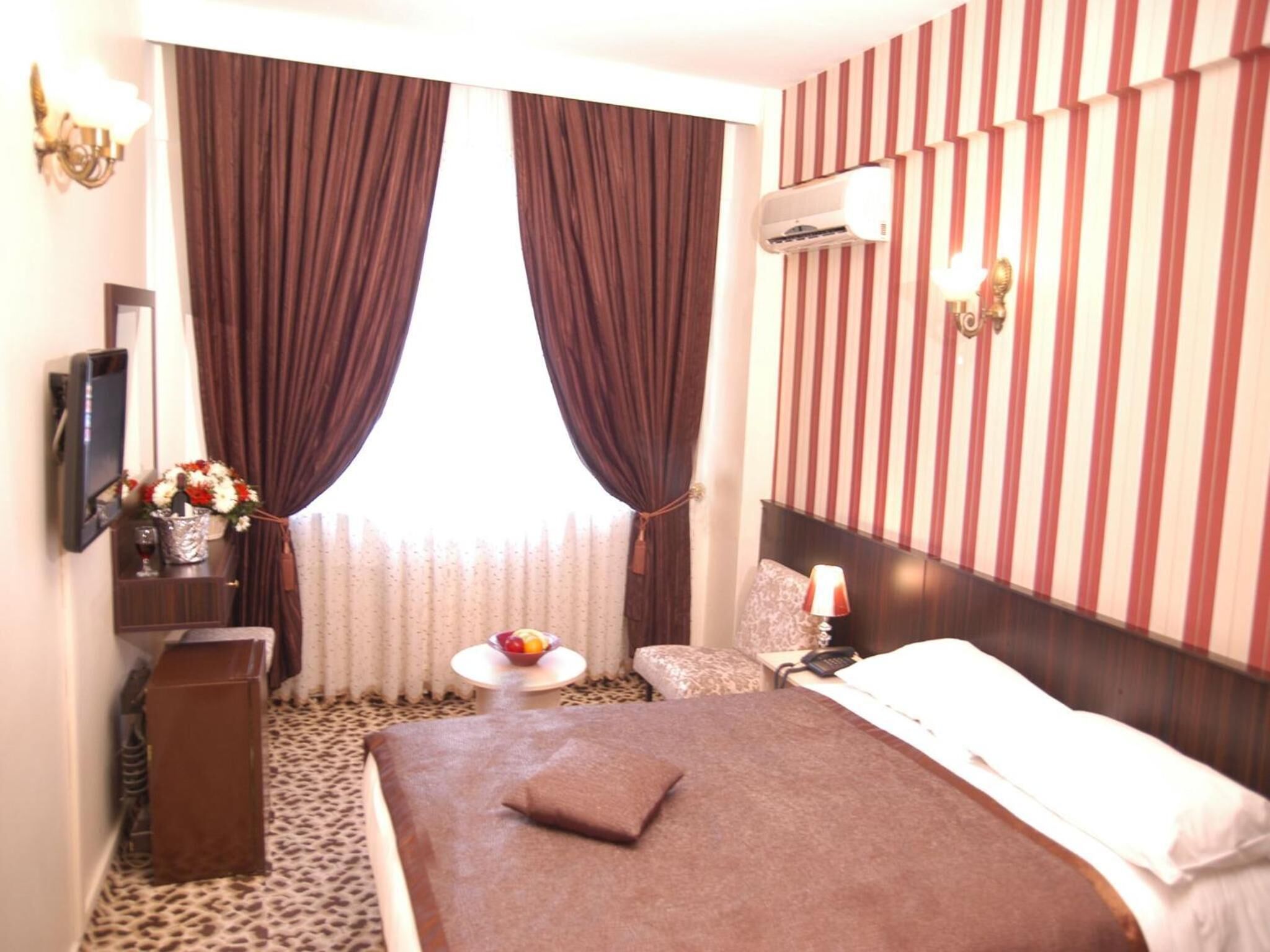 A hotel room with a large bed, a small round table with a bowl of fruit, a chair, a TV, and a wall-mounted air conditioner.