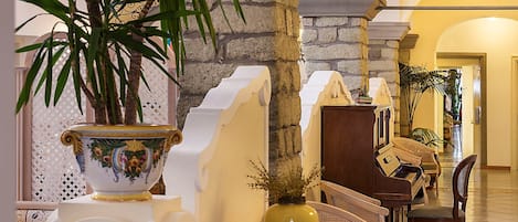 A lobby with a stone column, a large decorative vase, wicker chairs, and a piano.