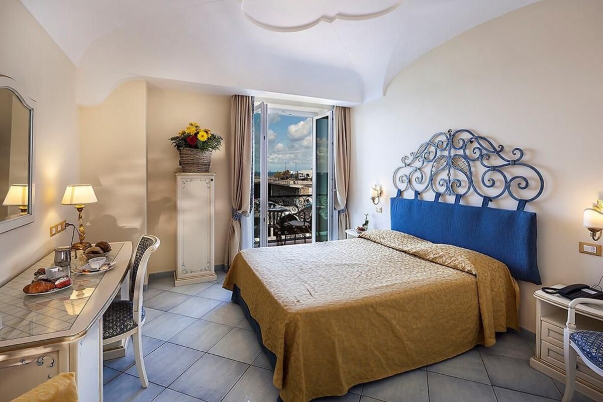 Una habitación de hotel con cama, escritorio con silla, pequeña mesa con lámpara, ventana con cortinas y cabecero decorativo de metal.