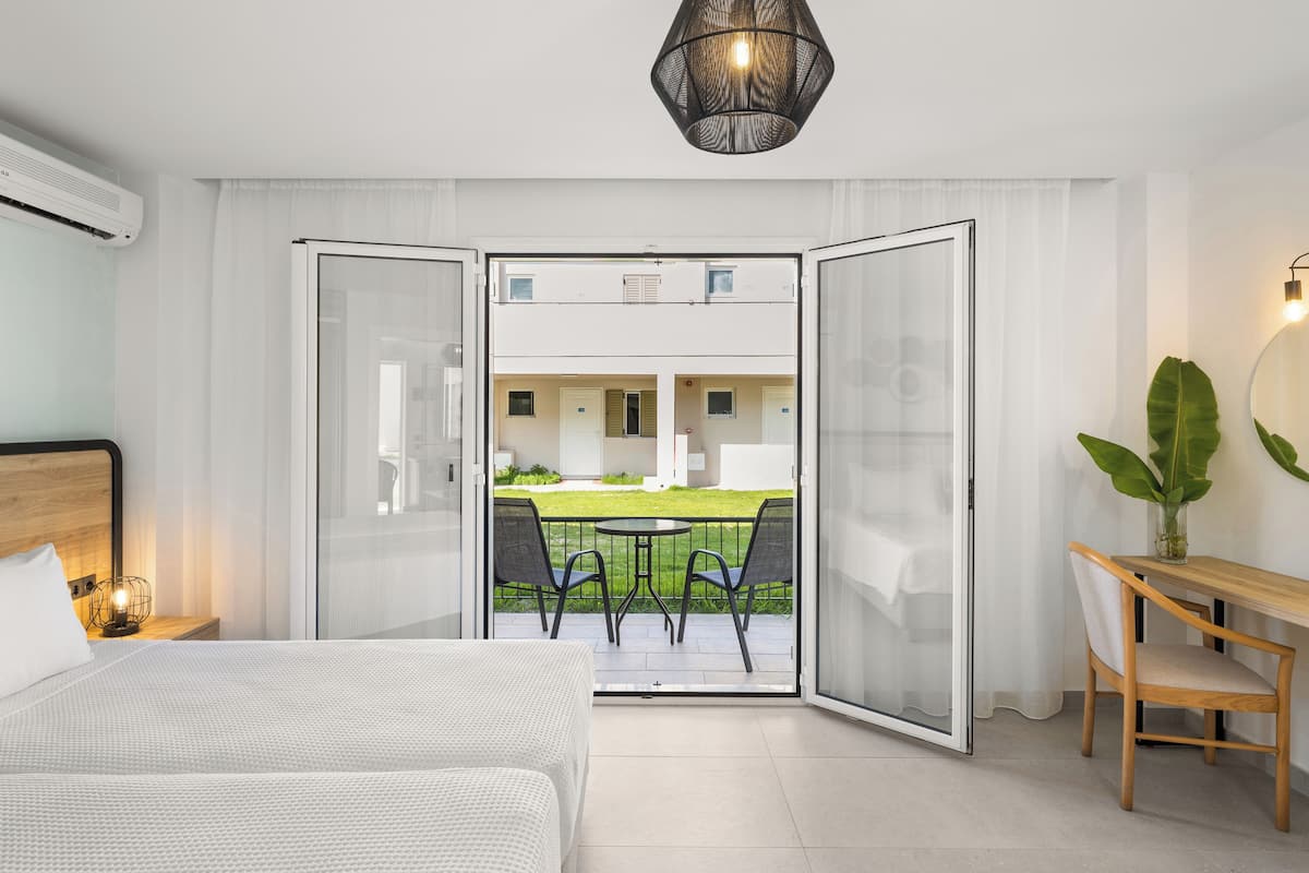 A hotel room with a bed, a desk with a chair, a table with chairs, and a view of a green lawn and building through a glass door.