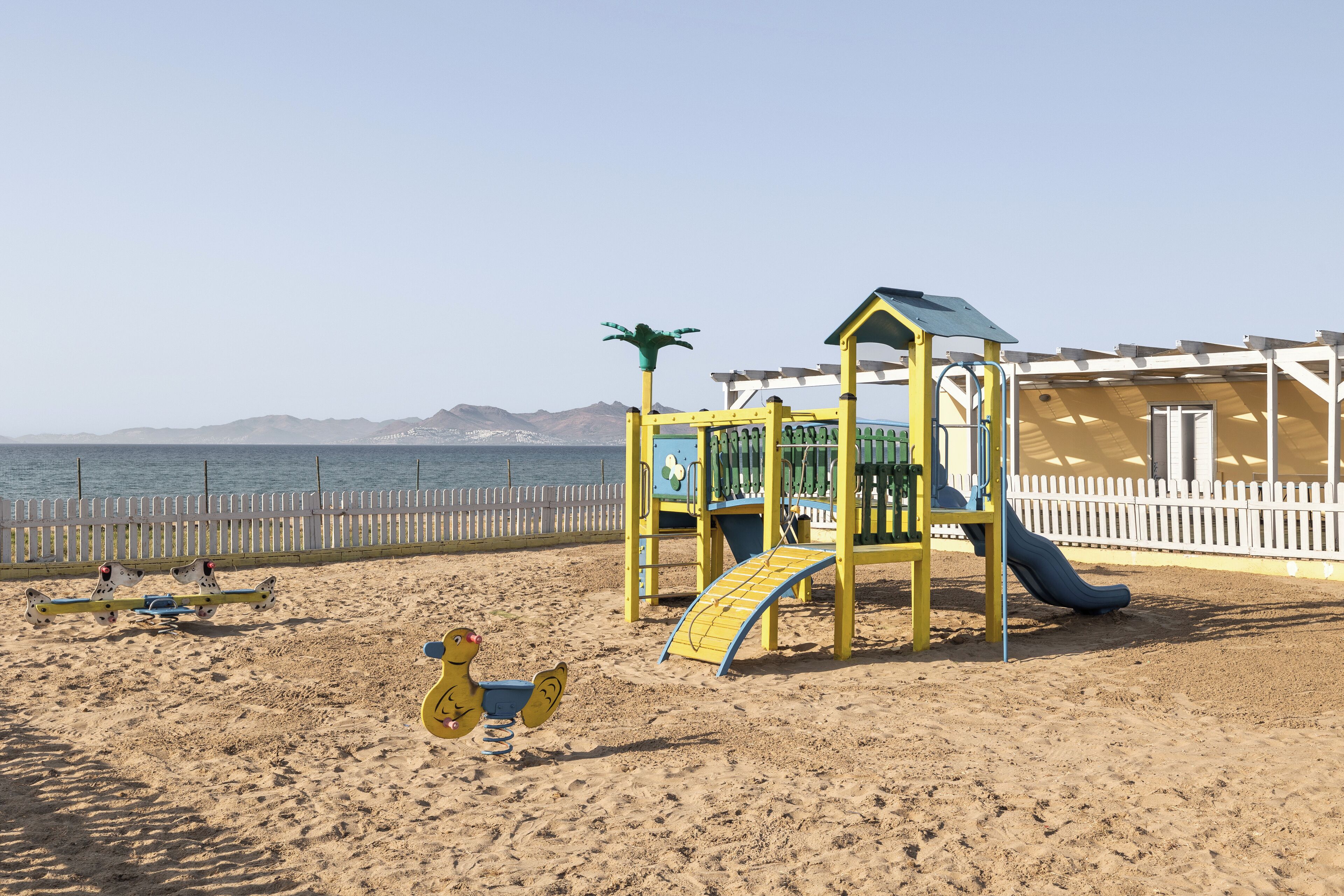A beach playground with a slide and swings, a fenced area, and a view of the sea and mountains in the background.