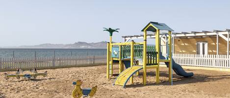 A beach playground with a slide and swings, a fenced area, and a view of the sea and mountains in the background.