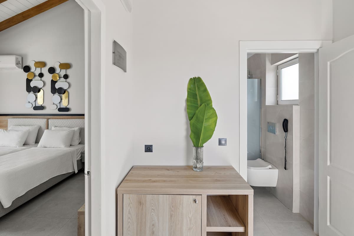 A modern bedroom with a bed, a wooden nightstand, a large green plant, and a bathroom visible through an open door.