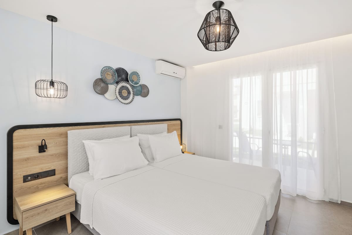 A modern bedroom with a wooden headboard, white bedding, a bedside table with a drawer, a hanging light fixture, and a decorative wall piece.
