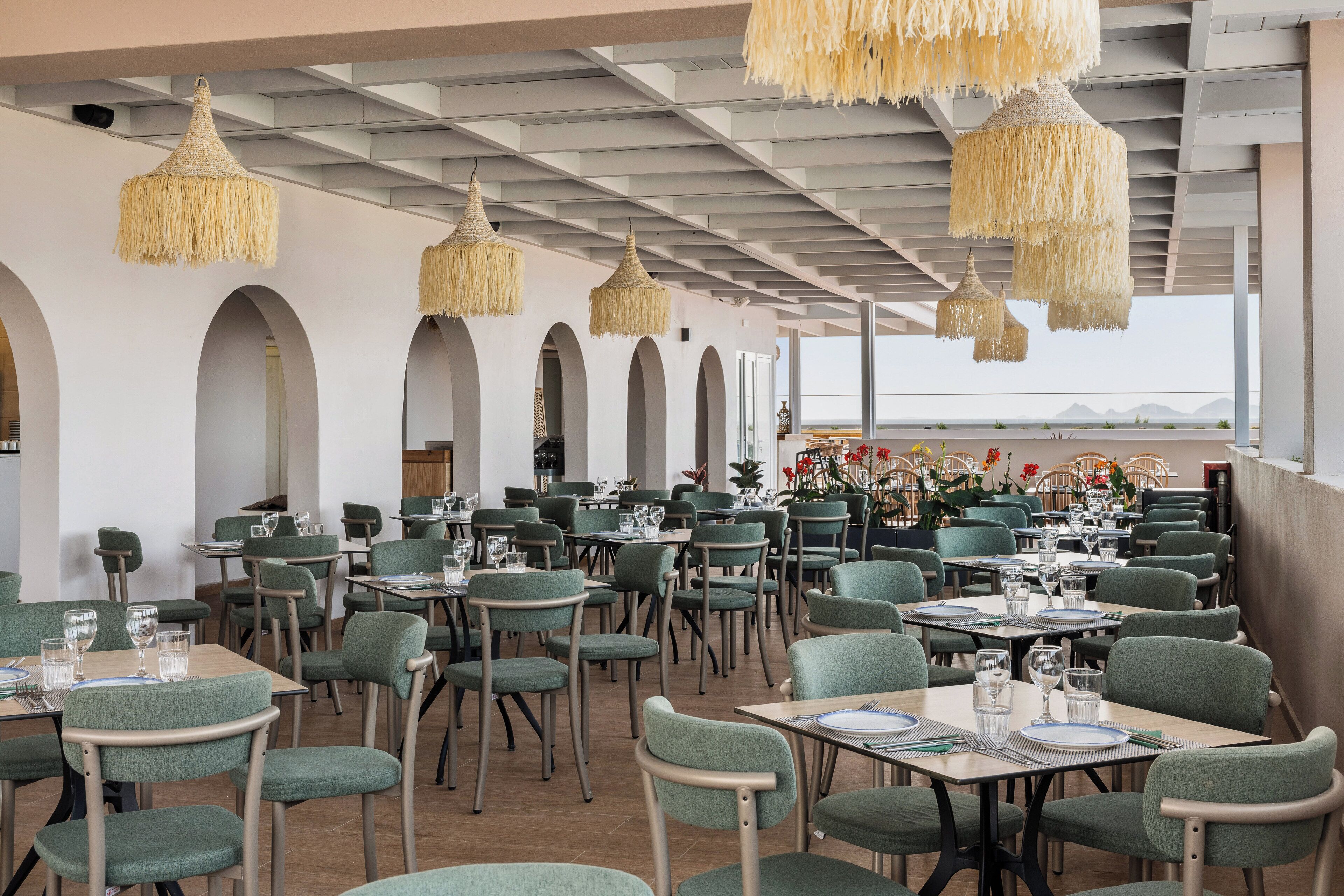 Un restaurante con sillas y mesas tapizadas de verde, lámparas colgantes tejidas y vistas al mar.