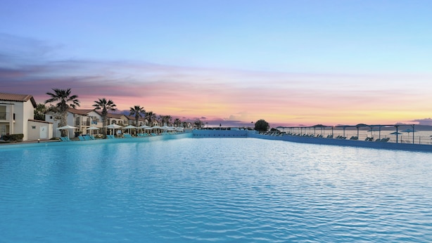 Una gran piscina con sillas de descanso y sombrillas, rodeada de edificios con balcones y palmeras.