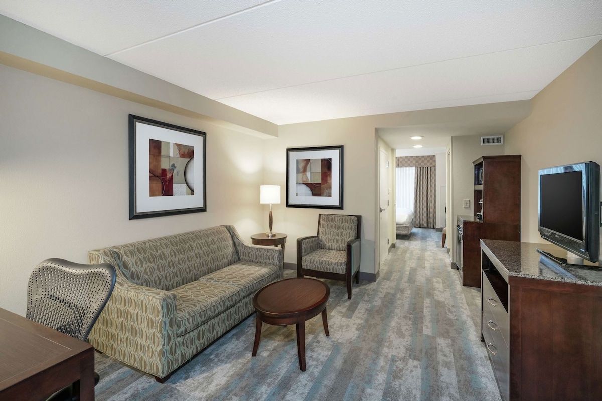 A hotel room with a patterned sofa, a small round table, a chair, a TV on a wooden stand, and framed artwork on the walls.