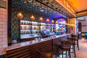 Zona de bar con mostrador, estanterías llenas de botellas, lámparas colgantes y taburetes.