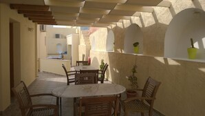 A covered outdoor patio with a round table and wooden chairs, surrounded by potted plants and a view of buildings in the background. - Anemomilos Suites (Santorini)