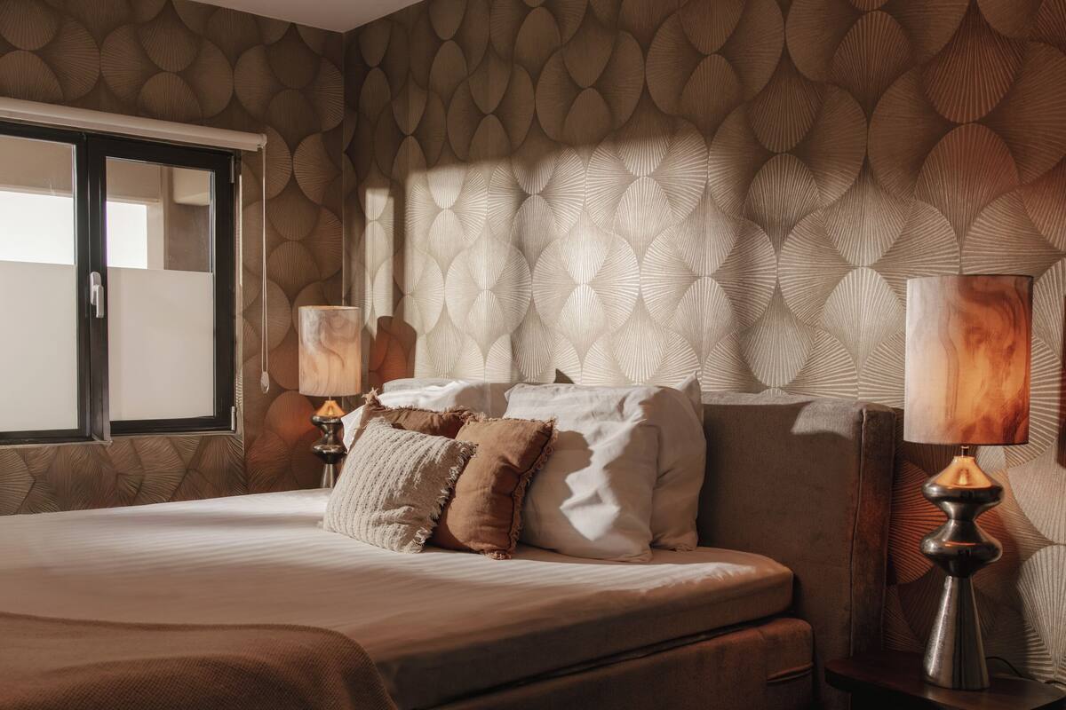 A bedroom with a large bed, two bedside lamps, a window, and a patterned wall.