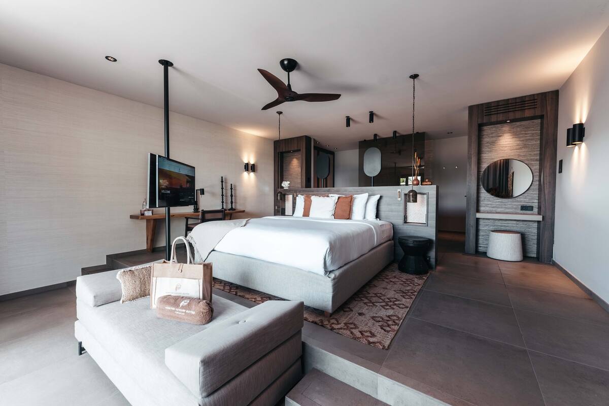 A modern bedroom with a large bed, a seating area, a TV, and a ceiling fan.