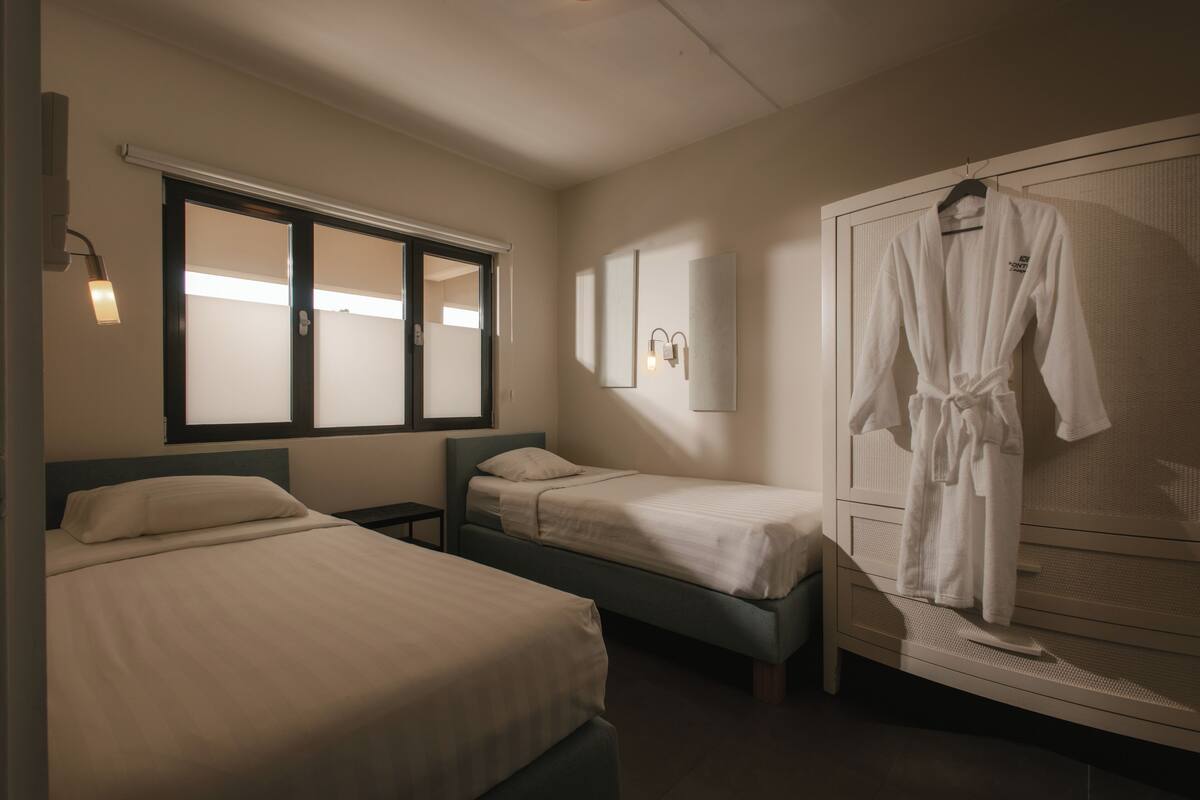 A hotel room with two single beds, a large window, a robe hanging on the wall, and a bedside table.