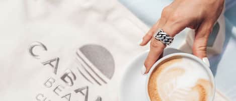A person holding a cup of coffee with a latte art design, a beach-themed tote bag, and a bracelet.