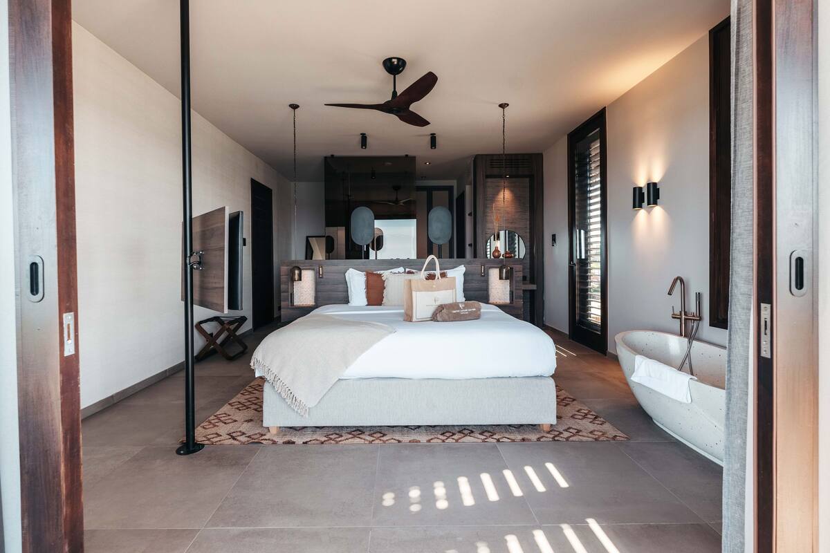 A modern bedroom with a large bed, a ceiling fan, a built-in bathtub, and a large mirror.