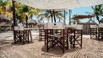 Mesas y sillas de madera al aire libre con letreros que dicen 'CABANA BEACH', sobre arena, con palmeras al fondo.