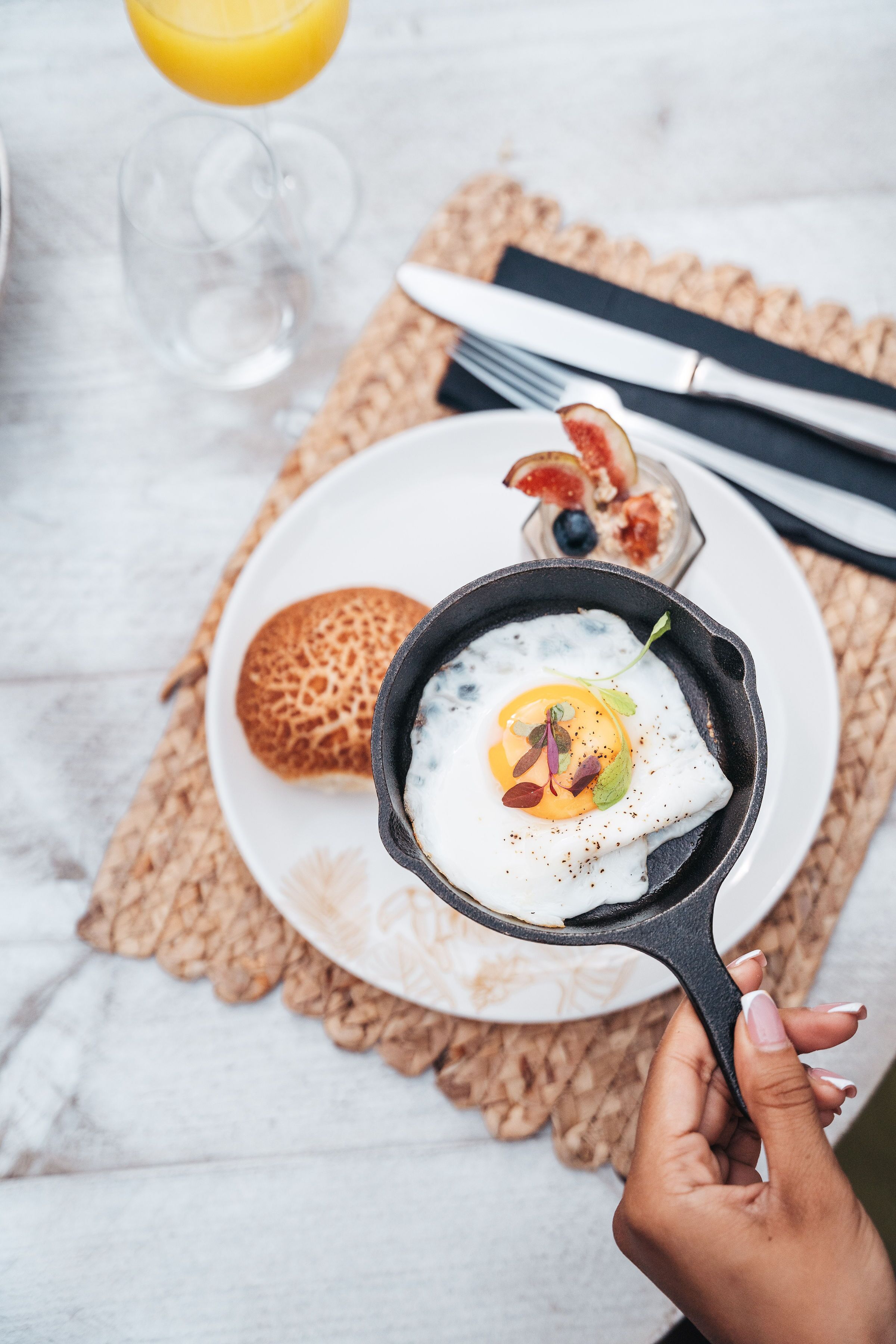 A breakfast setting with a skillet containing a poached egg, a plate with a bread roll, and a glass of orange juice.