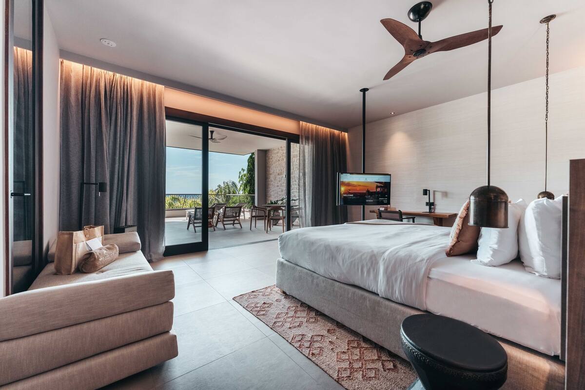 A modern bedroom with a large bed, a sofa, and a TV. There is a balcony with outdoor furniture.