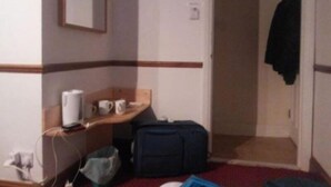 A hotel room with a wooden desk, a mirror, a bed, a suitcase, and a coat hanging on the wall. - Stansted Skyline Hotel (Dunmow)