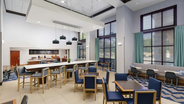 A modern dining area with a long counter, multiple tables, and chairs. There is a kitchen area with a counter and various items on display. - Hampton Inn & Suites West Sacramento (West Sacramento)