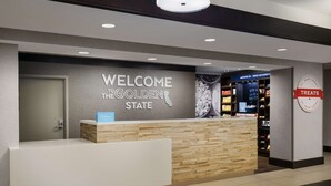 A reception desk with a "Welcome to the Golden State" sign, a blue Hilton sign, and a "Treats" sign on the wall. - Hampton Inn & Suites West Sacramento (West Sacramento)
