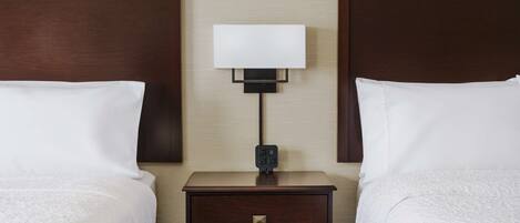 A hotel room with two beds, a nightstand with a lamp, and a wall-mounted electrical outlet.
