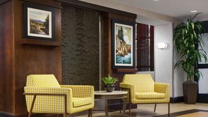 A lobby with two yellow patterned armchairs, a small round table with a plant, framed artwork on the walls, and a large potted plant. - Hampton Inn & Suites West Sacramento (West Sacramento)