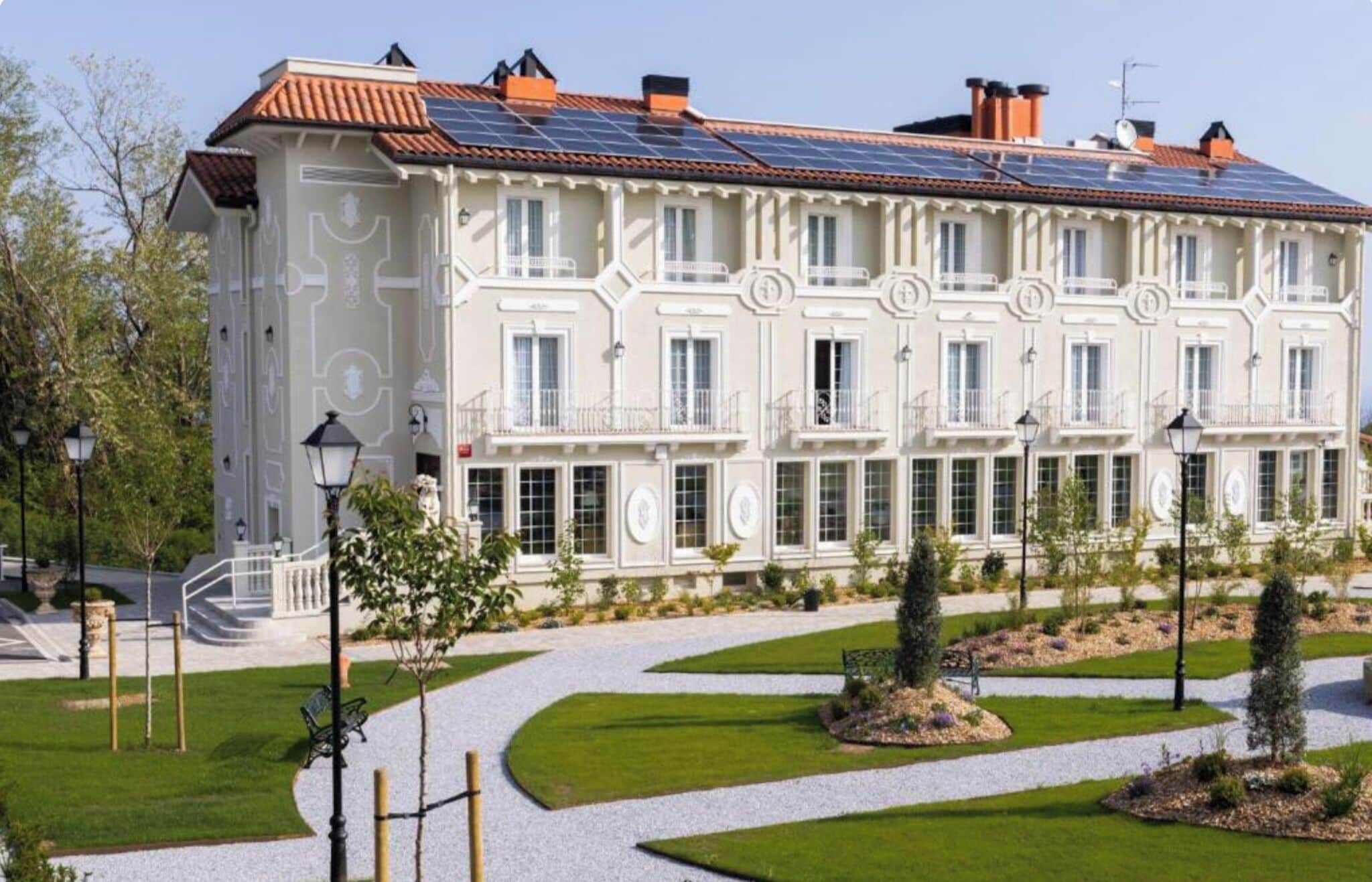 Un edificio grande de varios pisos con fachada simétrica, paneles solares en el techo y un jardín cuidado con senderos y árboles jóvenes.