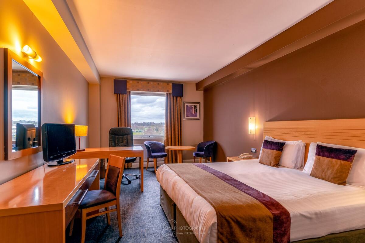 A hotel room with a bed, a desk with a TV, two chairs, a small table, and a window with curtains.