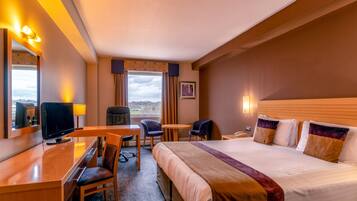 A hotel room with a bed, a desk with a TV, two chairs, a small table, and a window with curtains.