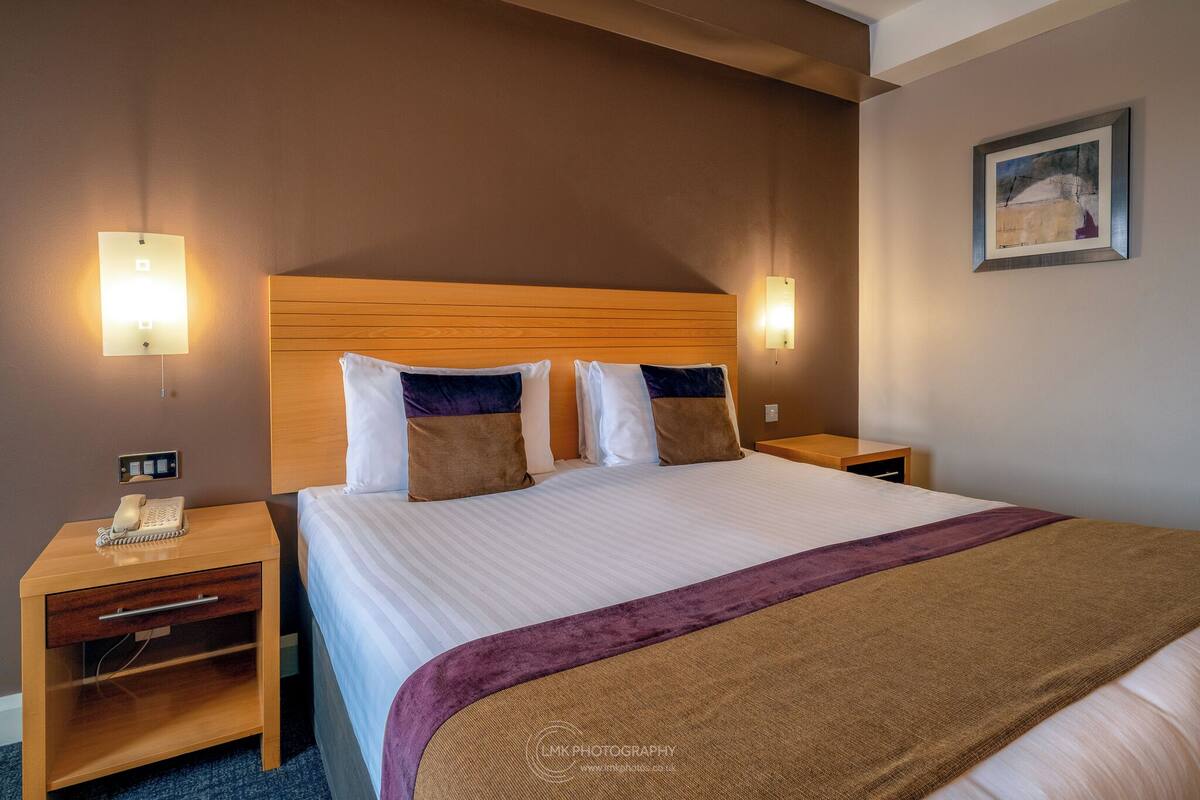 A hotel room with a large bed, two bedside tables, a nightstand, a telephone, and a painting on the wall.