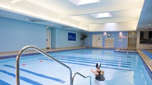 Piscina com faixas demarcadas, escada de metal e parede azul com relógio e placas.