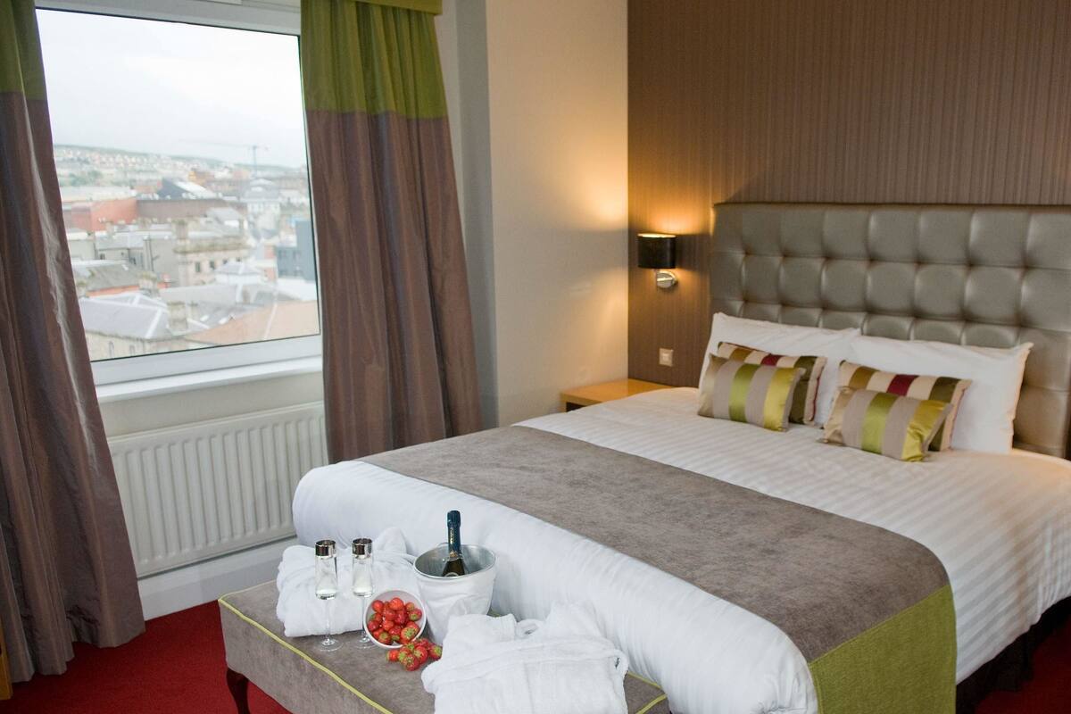 A hotel room with a large bed, a bedside table with a bottle and glasses, a tray with towels and fruit, and a view of the city through the window.