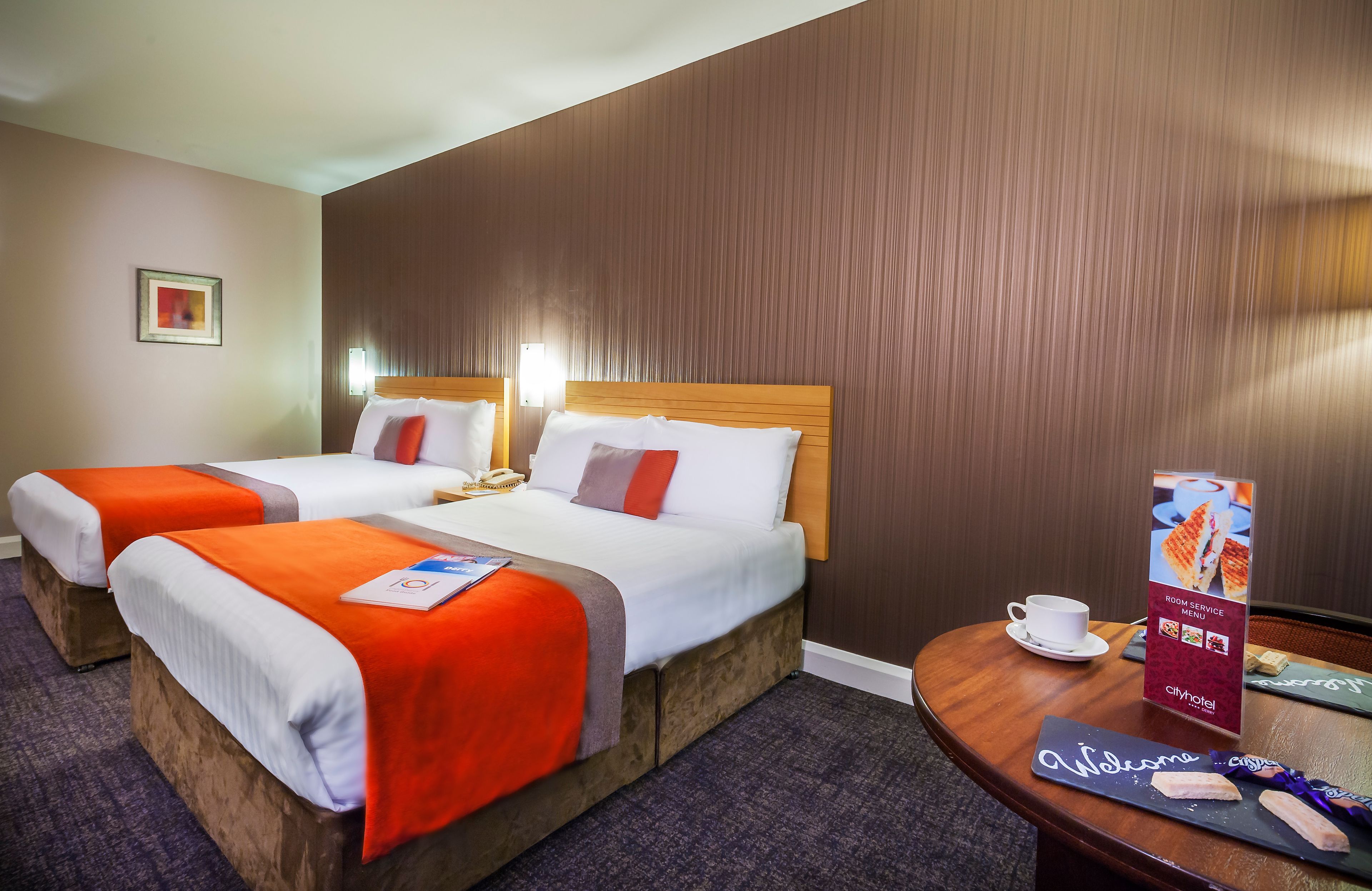 Ein Hotelzimmer mit zwei Betten, einem Holzkopfstück, einem kleinen Tisch mit Tasse und einem Willkommensschild, sowie einem an der Wand befestigten Schild.