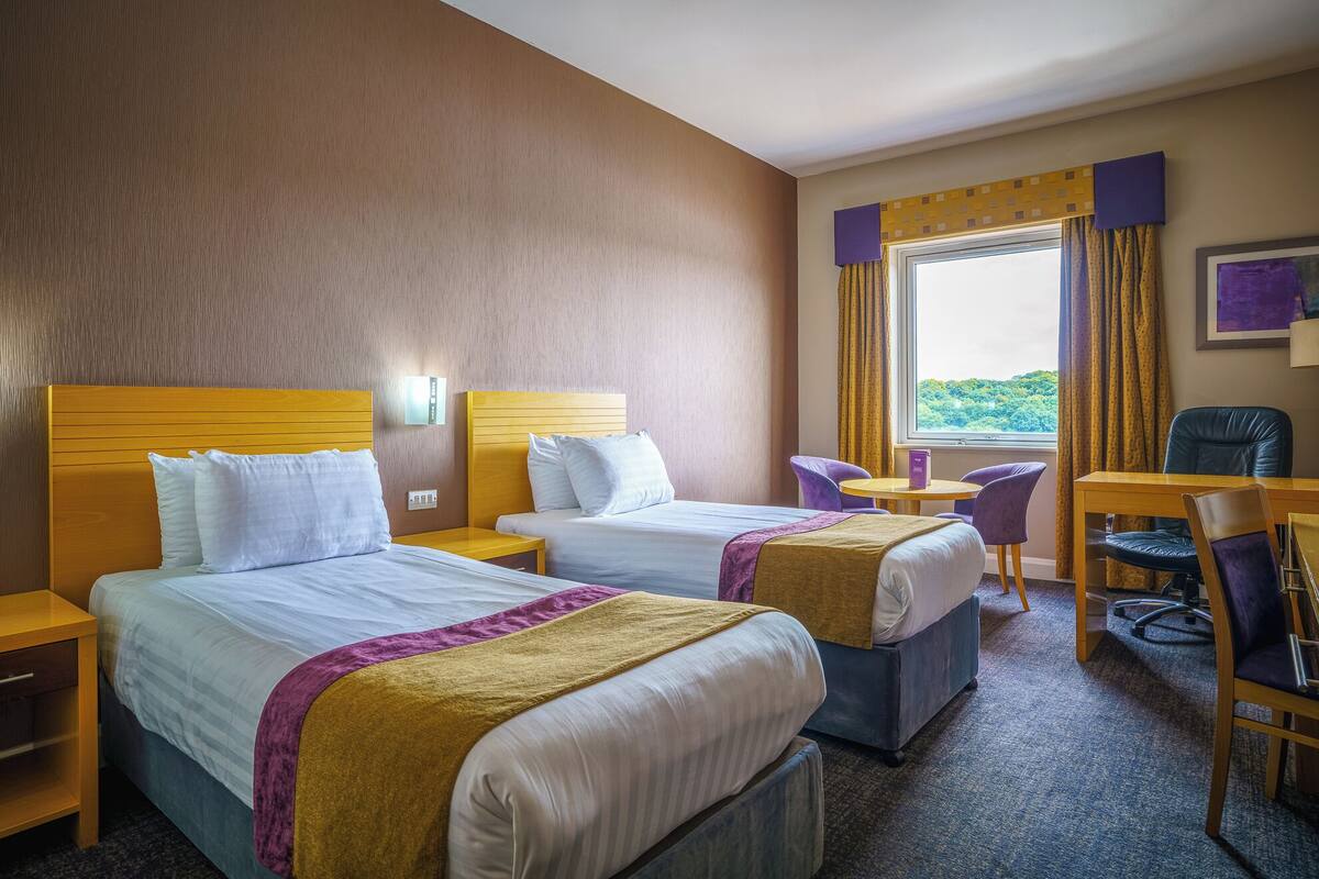 A hotel room with two beds, a desk, a chair, and a window with a view.