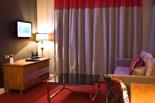 A hotel room with a wooden dresser, a desk with a lamp, a television on the wall, a sofa, and a large window with curtains.