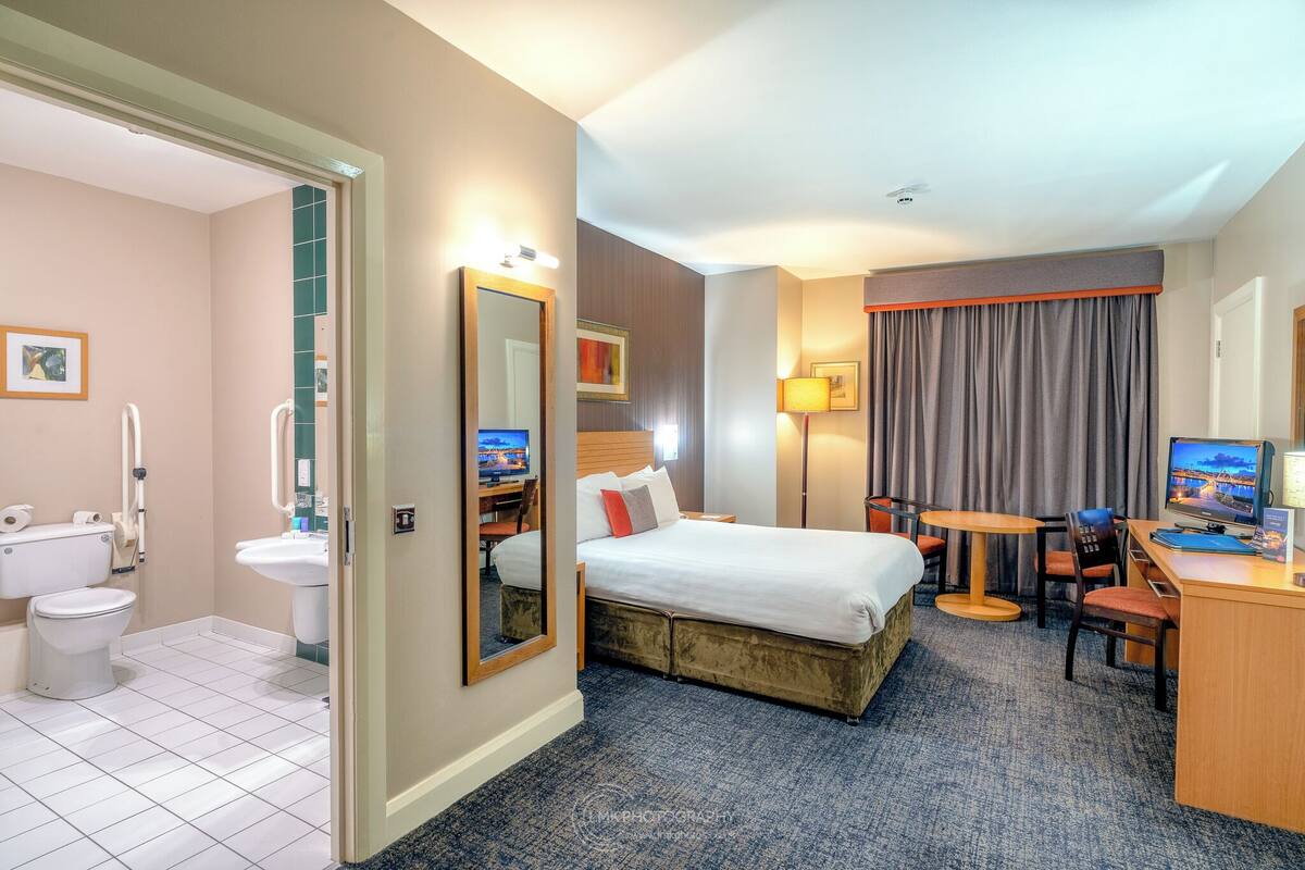 A hotel room with a bed, desk, chair, and a bathroom with a toilet and sink.