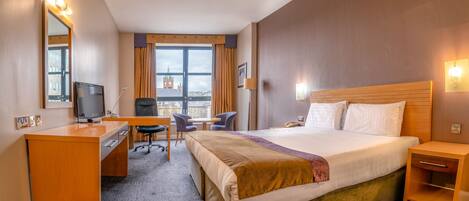 A hotel room with a bed, desk, chair, television, and a view of a cityscape.