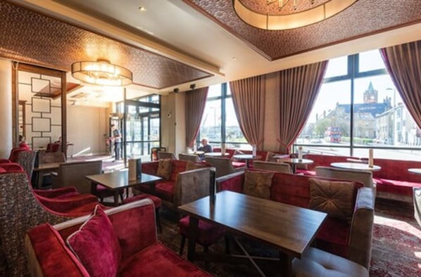 A restaurant with red upholstered seating, a patterned ceiling, and large windows offering a view of a cityscape. - City Hotel Derry (Londonderry)