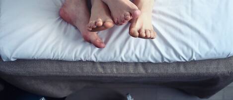 A bed with a white sheet and a grey mattress. There are two pairs of shoes on the floor, one pair is black with white soles, and the other pair is blue with white soles.
