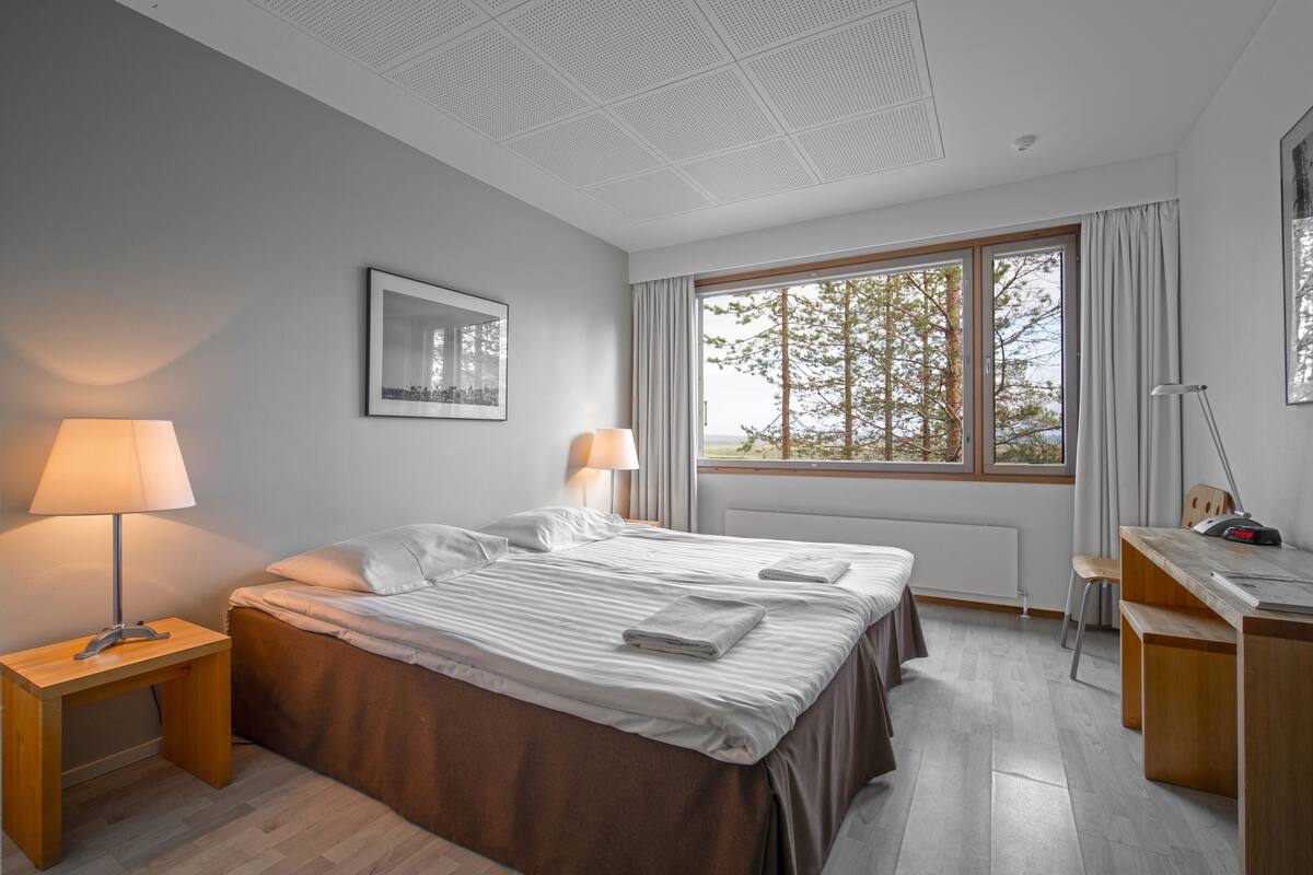 A modern hotel room with a large bed, a desk, and a window with a view of trees.