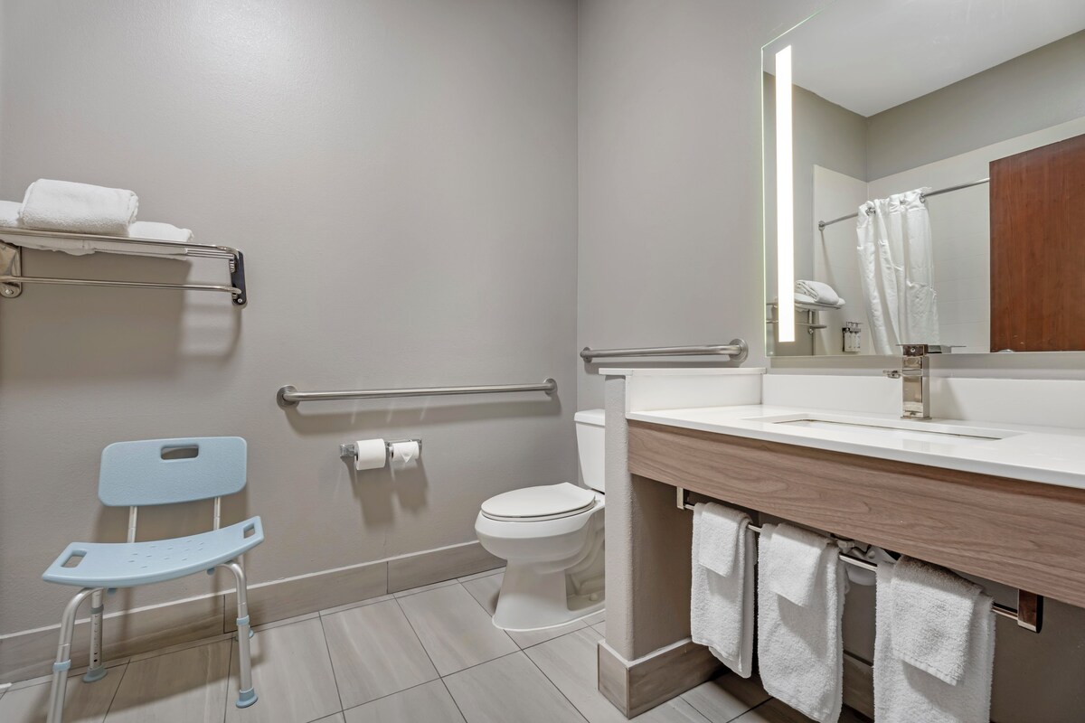 A modern bathroom with a toilet, sink, mirror, and a wheelchair-accessible chair.
