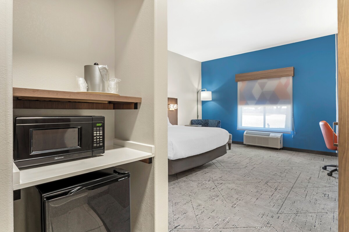 A hotel room with a microwave, a small refrigerator, a bed, a window with blinds, a desk lamp, and a chair.