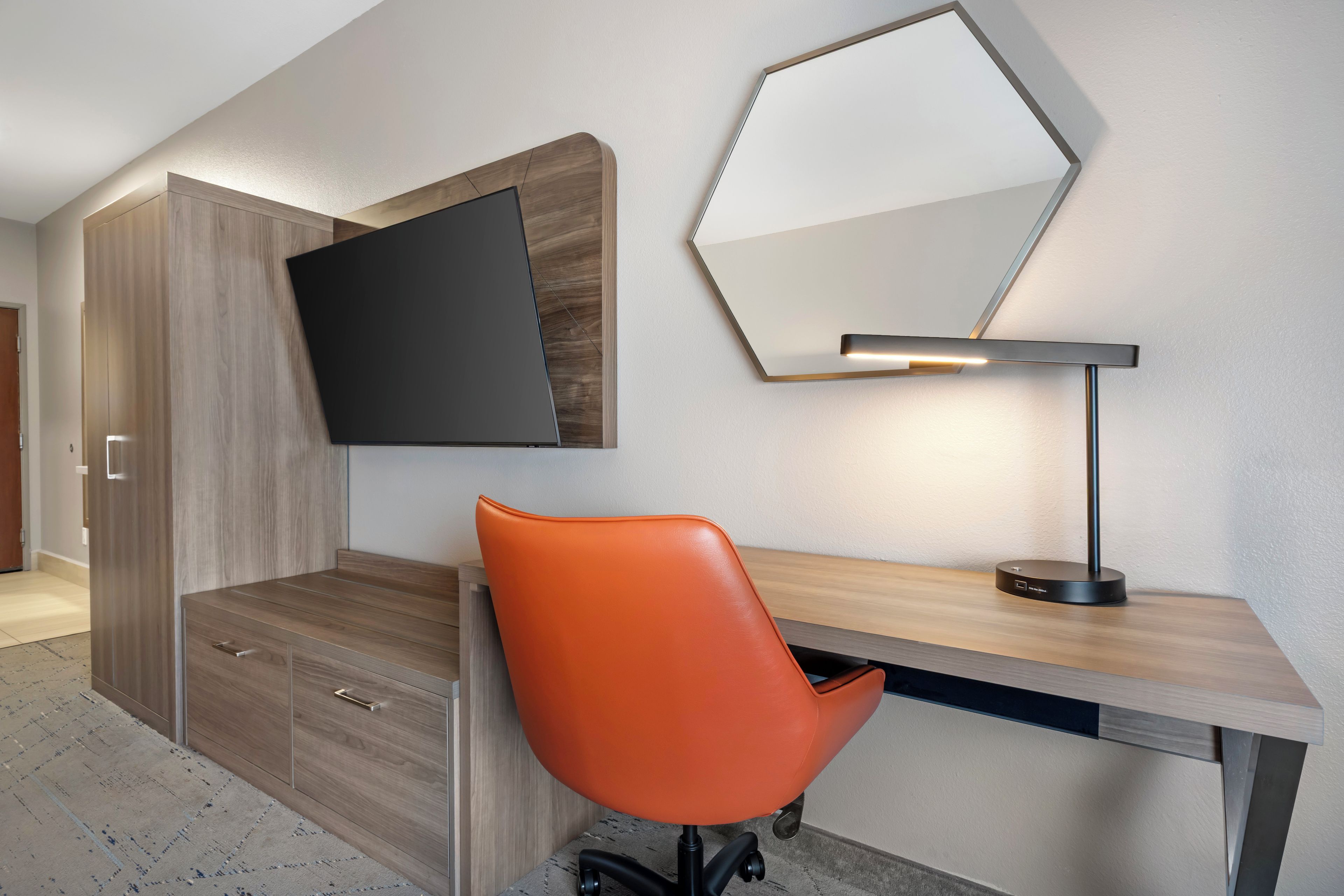 Un espace de bureau moderne comprenant un bureau, une chaise de bureau orange, un téléviseur fixé au mur et une lampe de bureau.