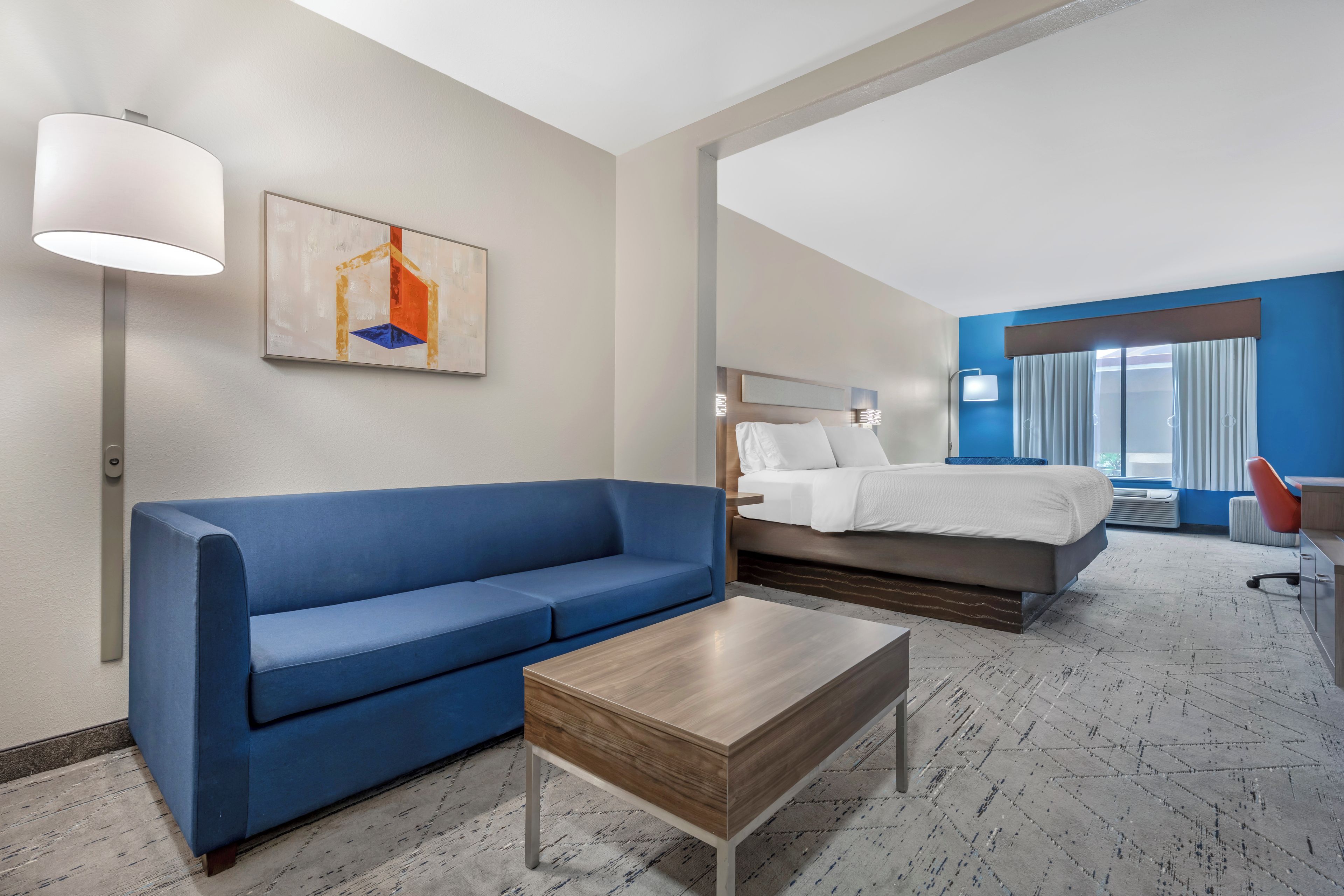 A hotel room with a blue sofa, a wooden coffee table, a bed with white bedding, and a painting on the wall.