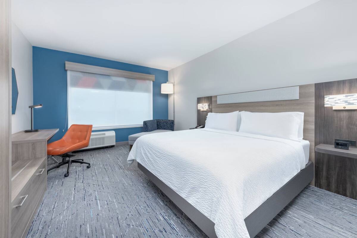 A modern hotel room with a large bed, a desk, an orange chair, a window with blinds, and a nightstand with a lamp.