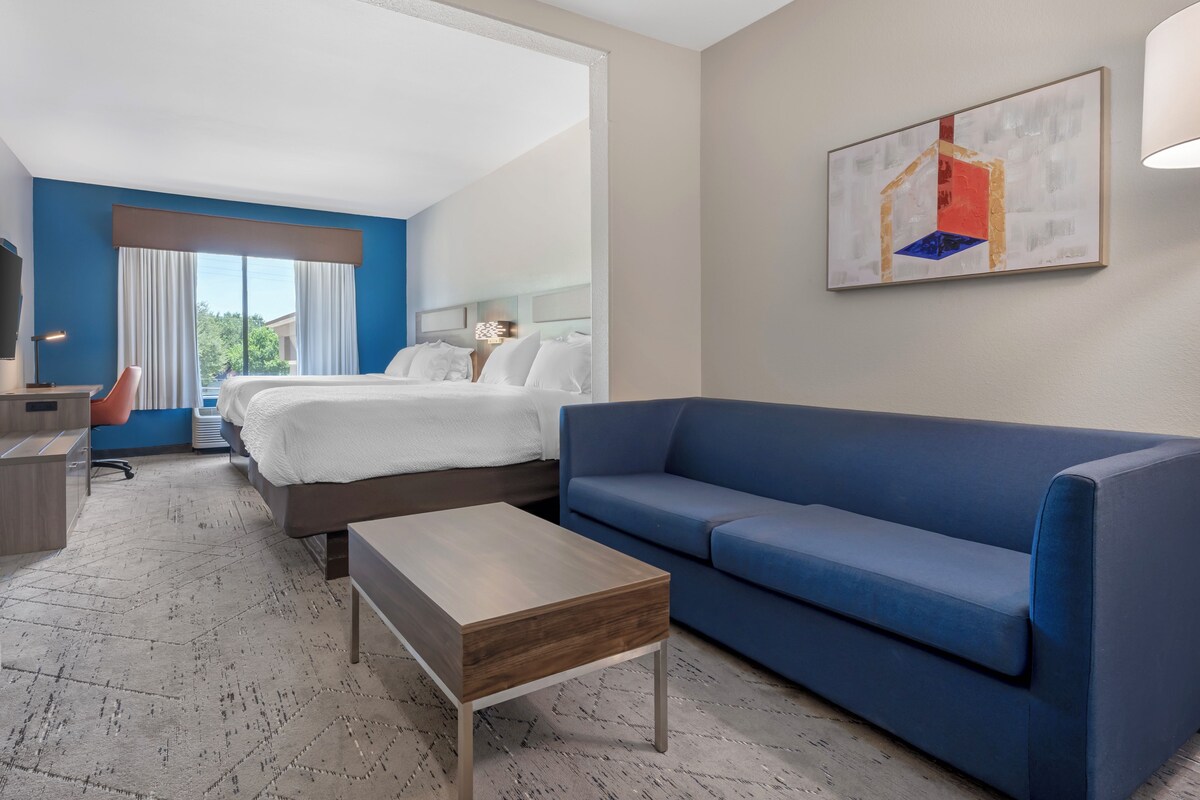 A hotel room with a bed, a blue sofa, a small wooden table, a desk with a chair, and a painting on the wall.